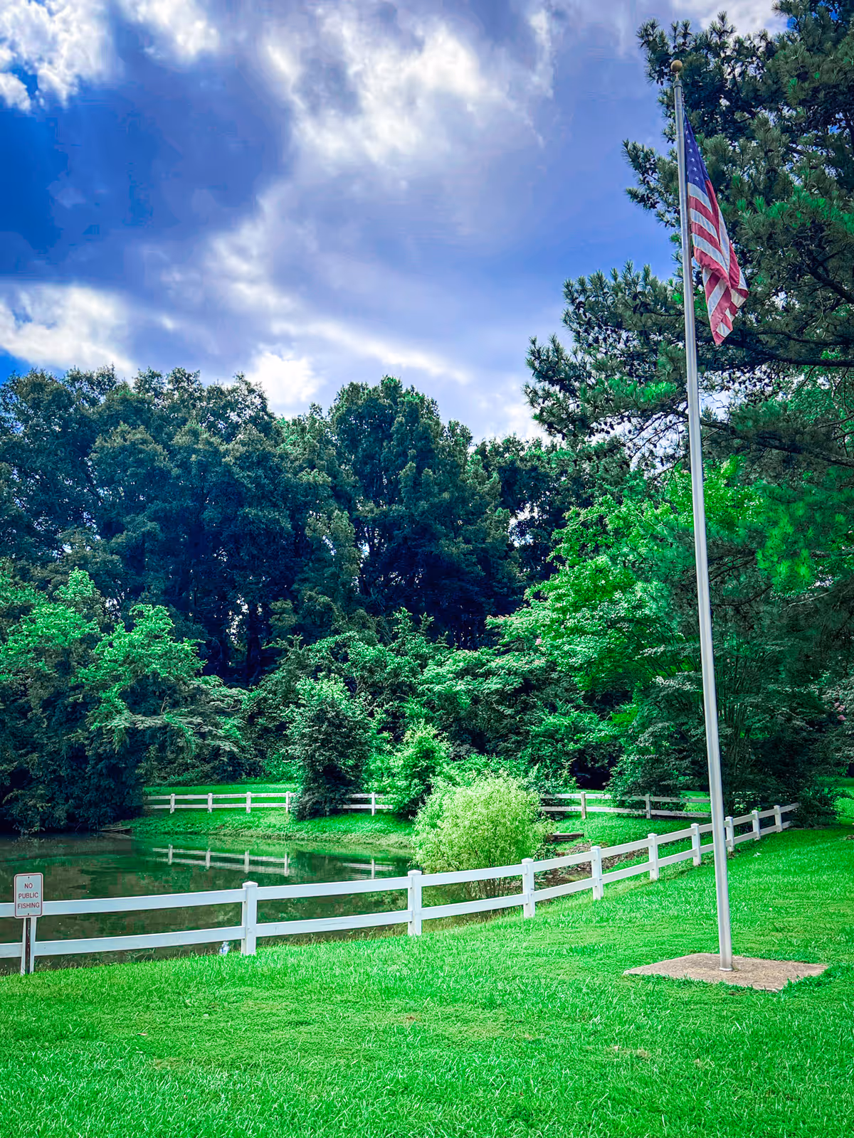A peaceful outdoor scene at Spring Terrace Assisted Living featuring a small pond surrounded by a white fence, lush green grass, and dense trees in the background under a partly cloudy sky. An American flag on a flagpole stands prominently on the right side of the image.