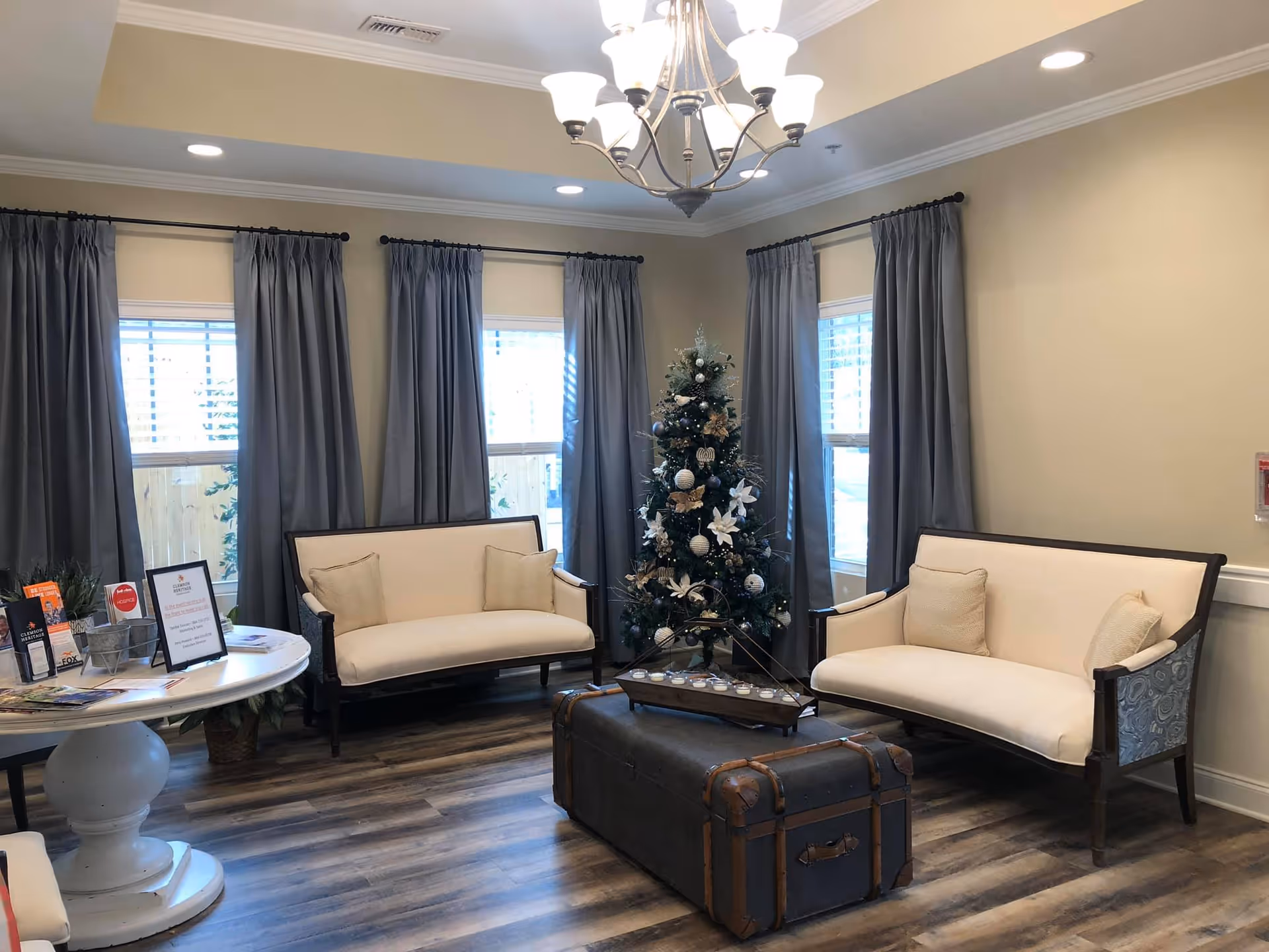 Light-filled seating area with two cream settees, a decorated Christmas tree, a trunk coffee table and a round brochure table in a senior living common room.