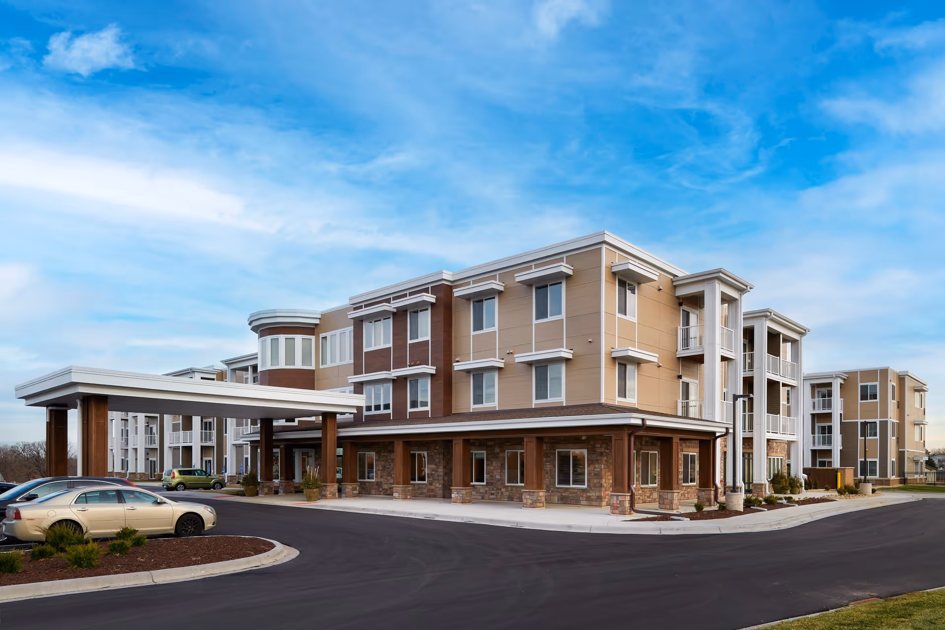 Exterior view of a modern three-story senior living facility building with a covered entrance, multiple windows, and a parking area with several cars. The building features beige and brown tones with stone accents and white trim under a partly cloudy blue sky.