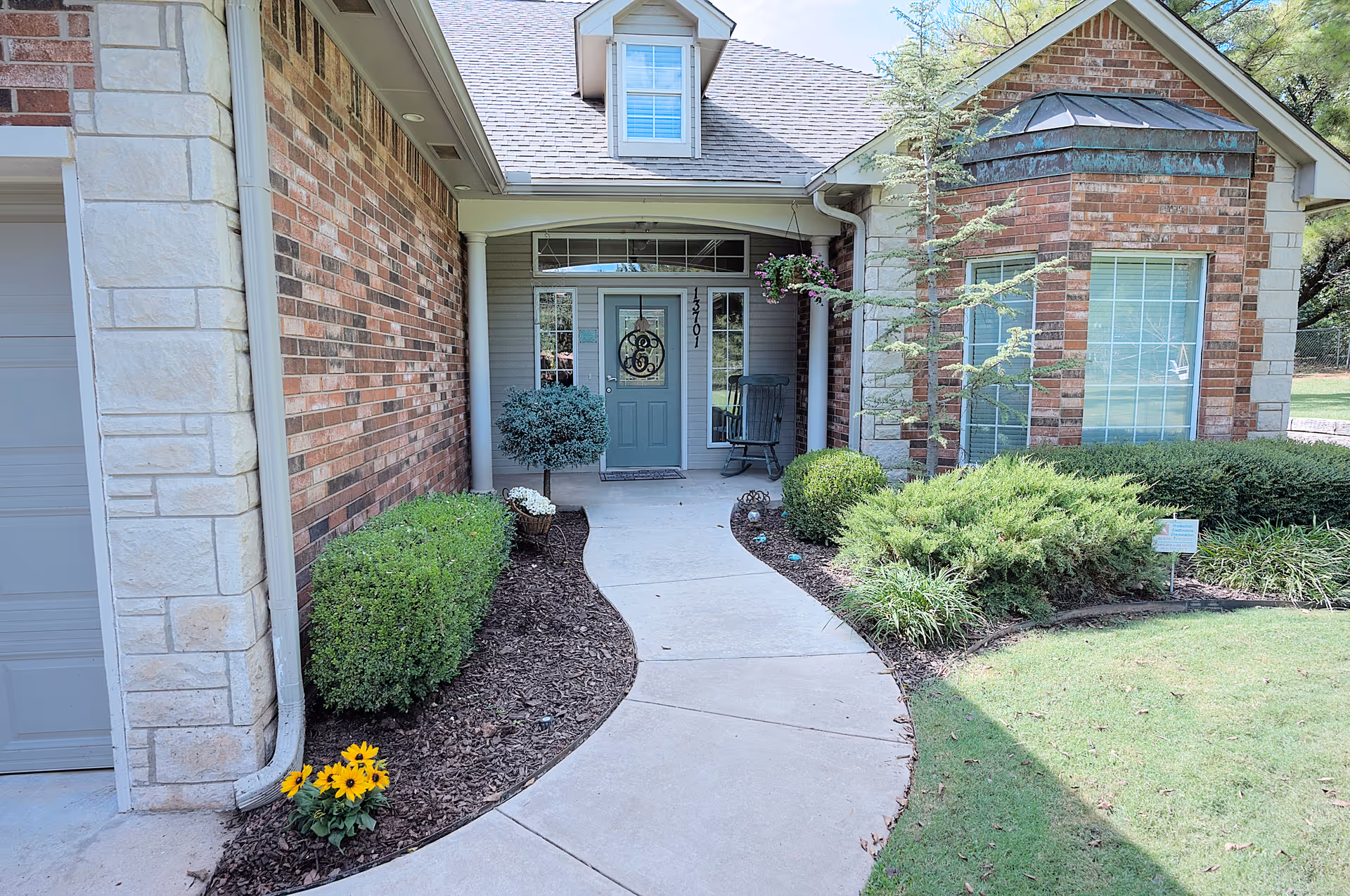 Front entrance of a brick and stone house with a curved concrete walkway leading to a green door with decorative glass. The entrance is flanked by bushes, small trees, and flower beds with mulch. There is a rocking chair on the porch and a hanging flower basket near the door.