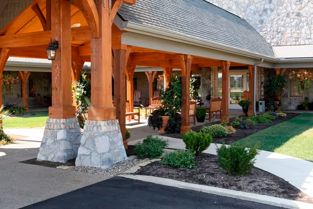 Covered outdoor seating area with wooden beams and stone bases at the entrance of a building, surrounded by landscaped plants and greenery.