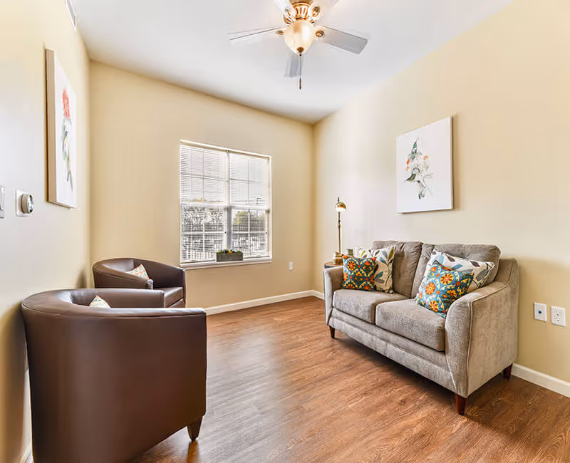 Bright living room with a gray loveseat, two brown armchairs, hardwood floors, a window, and a ceiling fan.