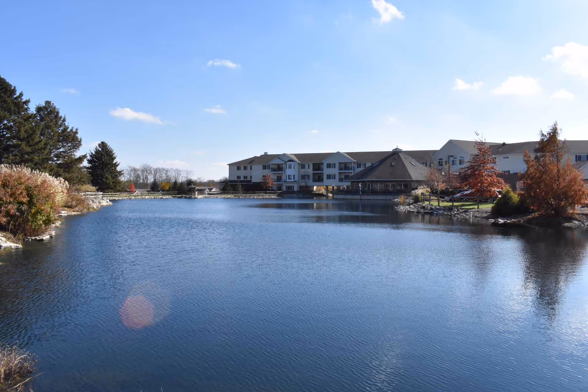 A large pond with calm water in the foreground, surrounded by trees with autumn foliage and a multi-story building in the background under a blue sky with a few clouds.