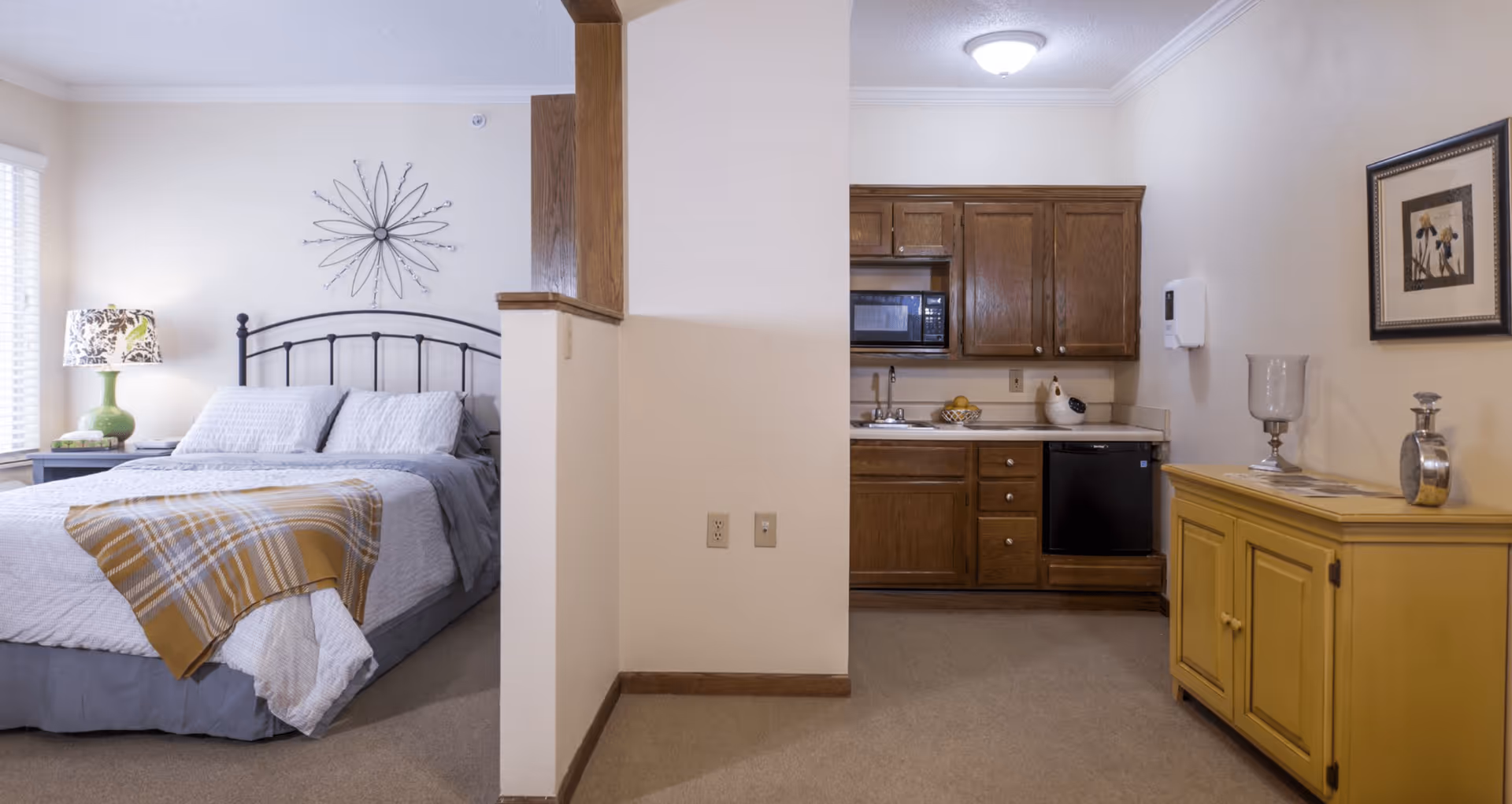 Interior view of a senior living facility room showing a bedroom area with a bed, pillows, a bedside table with a lamp, and a decorative wall piece. Adjacent to the bedroom is a small kitchen area with wooden cabinets, a microwave, a sink, a mini refrigerator, and a yellow cabinet with decorative items on top.