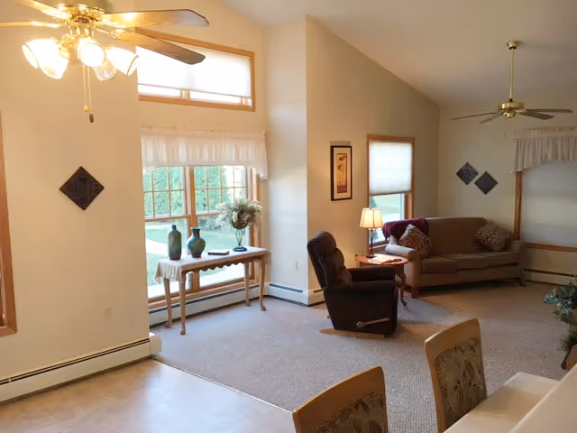 A cozy living room area with a beige sofa, a dark recliner chair, a small side table with a lamp, and a wooden table with decorative vases and a flower arrangement near a large window. The room has carpeted flooring, ceiling fans with lights, and neutral-colored walls with framed artwork and window treatments.