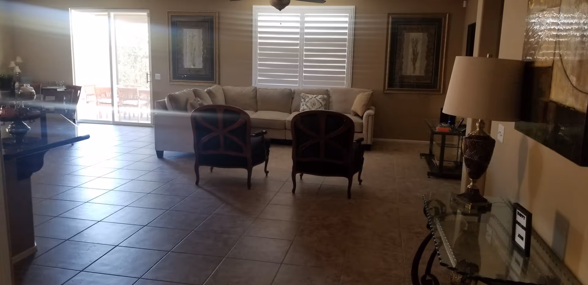 A living room with a beige sectional sofa, two dark wooden chairs with cushions, and two framed artworks on the wall. There is a glass sliding door leading to an outdoor patio with chairs. The room has tiled flooring, a glass table with a decorative lamp, and a ceiling fan above.