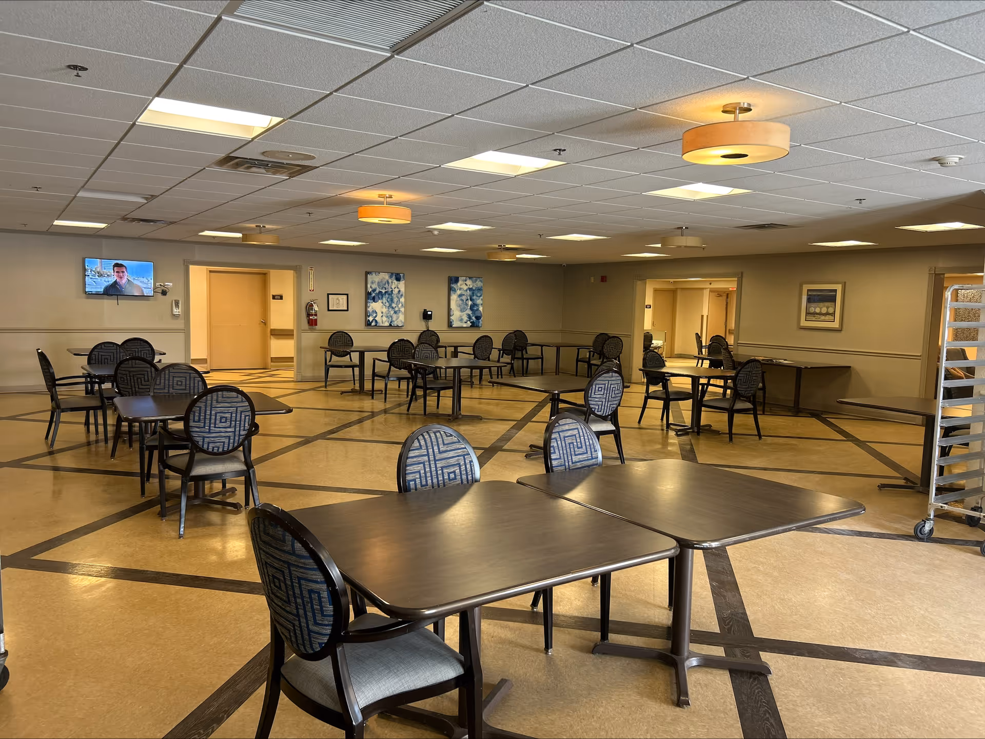 A spacious dining room with several dark wooden tables and patterned cushioned chairs arranged neatly. The room has a beige floor with dark geometric patterns, beige walls adorned with framed artwork, and a ceiling with recessed lighting and hanging fixtures. A television is mounted on one wall, and there are open doorways leading to other areas.