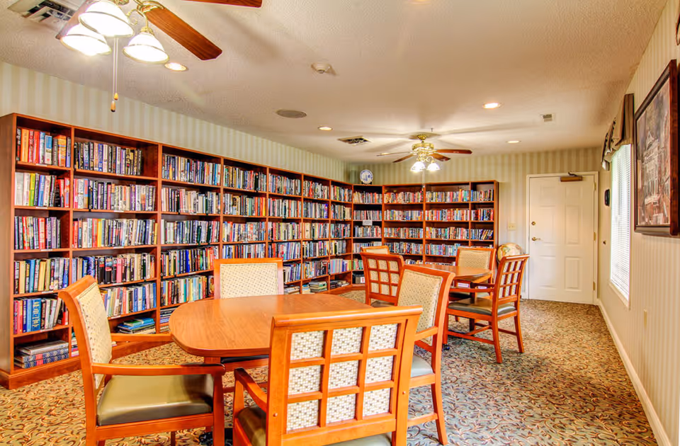 A cozy library room with wooden bookshelves filled with books along two walls. There are two wooden tables surrounded by cushioned chairs with patterned backs. The room has a patterned carpet, striped wallpaper, ceiling fans with lights, and a window with blinds on the right side. A door is visible at the far end of the room.