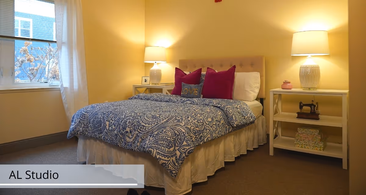 A cozy bedroom with a bed featuring a blue and white patterned comforter and three magenta pillows along with a small decorative pillow that says 'LOVE'. There are two white nightstands on either side of the bed, each with a white lamp. One nightstand has a small framed picture, and the other has a vintage sewing machine and decorative boxes. A window with sheer white curtains lets in natural light.