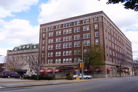 A large multi-story brick building with numerous windows and red awnings over the ground floor entrances, situated on a street corner with some parked cars and trees nearby under a partly cloudy sky.