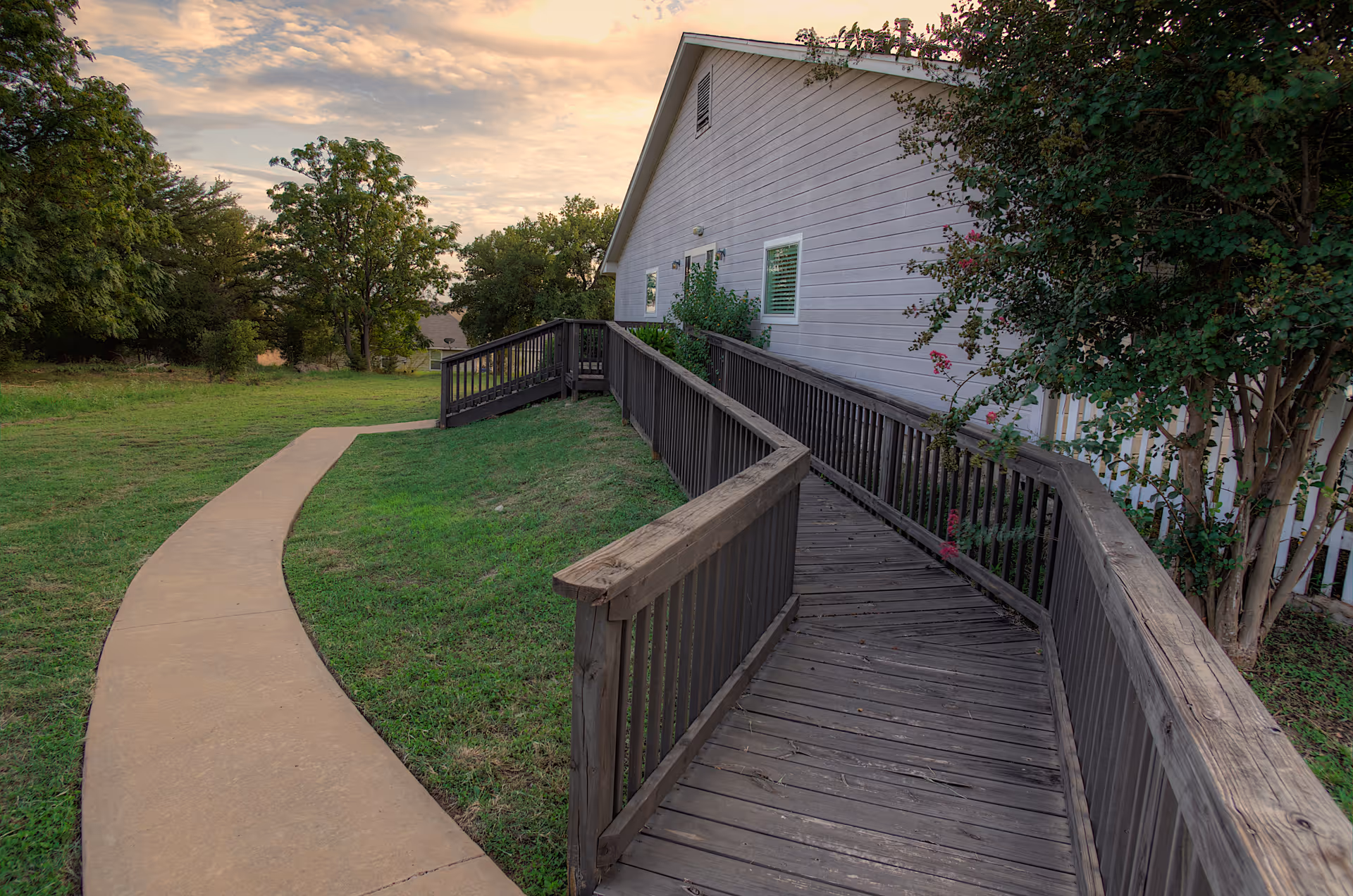 A wooden wheelchair-accessible ramp leading to the entrance of a light-colored building, surrounded by green grass, trees, and a curved concrete walkway under a partly cloudy sky during sunset.