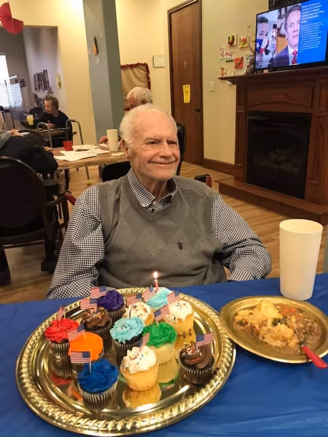 An elderly man sitting at a table with a blue tablecloth, smiling. In front of him is a gold tray with colorful cupcakes decorated with small American flags and one lit candle. Next to the tray is a plate with food and a white cup. In the background, other elderly people are seated at tables in a communal dining area with a TV mounted above a wooden fireplace.