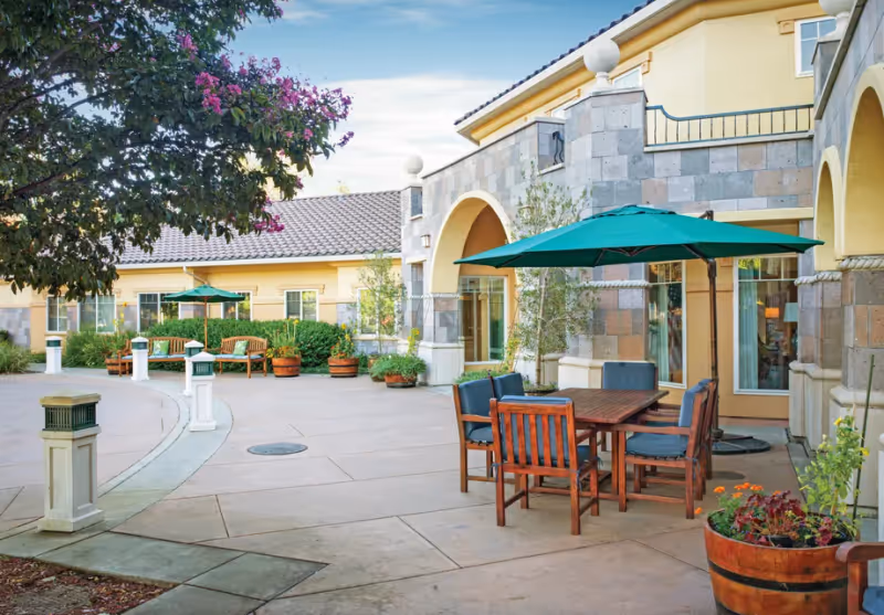 Outdoor patio area at Cogir of Sonoma Plaza featuring wooden tables and chairs with green umbrellas, potted plants, benches, and a building with stone and yellow walls under a partly cloudy sky.