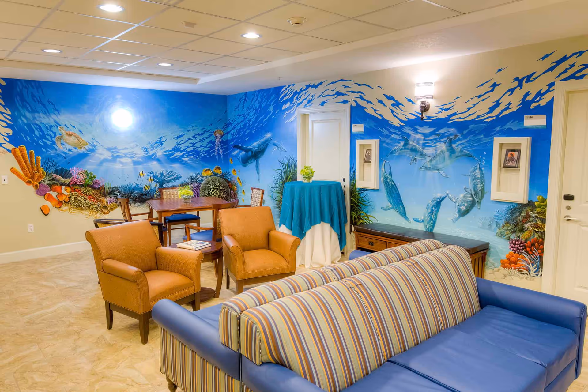 A cozy common area with a striped blue sofa, two orange armchairs, a wooden table with chairs, and a small round table covered with a blue cloth. The walls feature a vibrant underwater mural depicting sea turtles, fish, coral reefs, and dolphins swimming in the ocean.