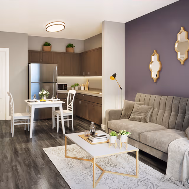 A modern living space featuring a gray sofa with cushions and a throw blanket, a white coffee table with books and small plants, and a dining area with a white table and two chairs. The kitchen area has dark wood cabinets, a stainless steel refrigerator, a microwave, and various kitchen items. The walls are painted in neutral tones with a purple accent wall decorated with two gold-framed mirrors. The floor is dark wood, and there is a white patterned rug under the coffee table.