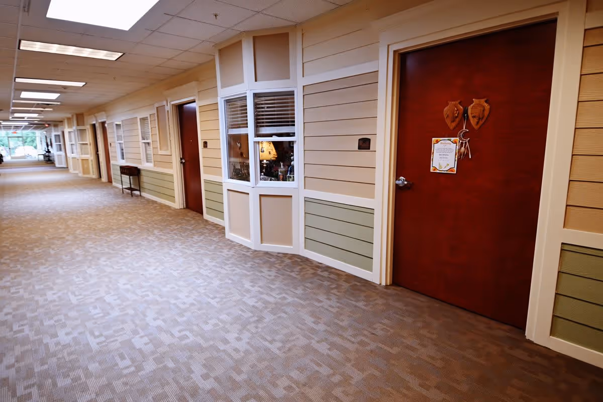 A long, carpeted hallway in a senior living facility with beige and green paneled walls, several closed wooden doors, and a window showing a room with a lamp inside. The ceiling has fluorescent lights and a fire sprinkler.