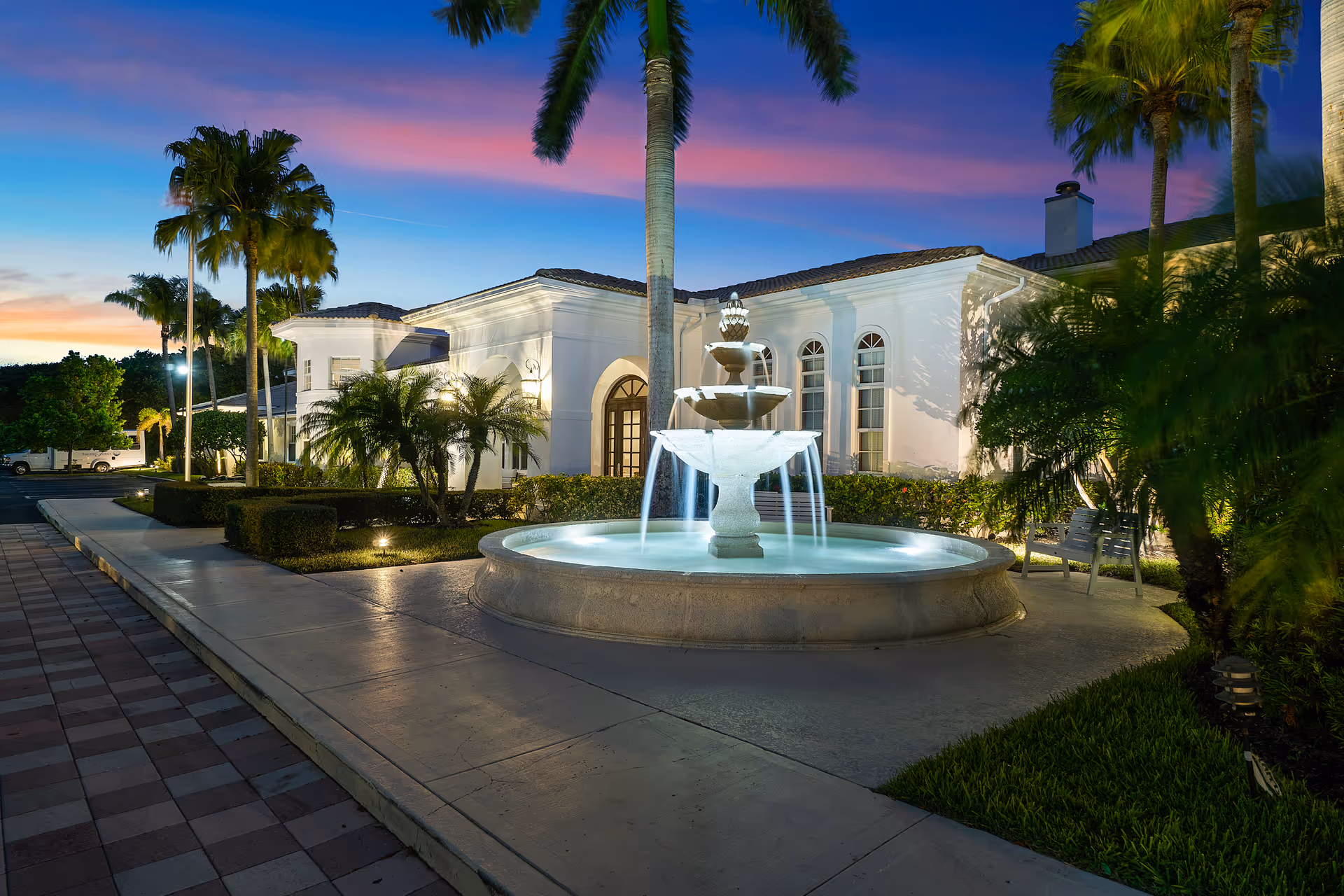 Front exterior of a white Mediterranean-style building with a lit central fountain, palm trees, and a colorful dusk sky.