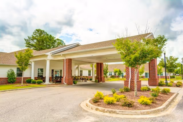 Entrance area of a senior living facility with a covered drop-off zone supported by brick columns, surrounded by small trees and landscaped greenery under a partly cloudy sky.