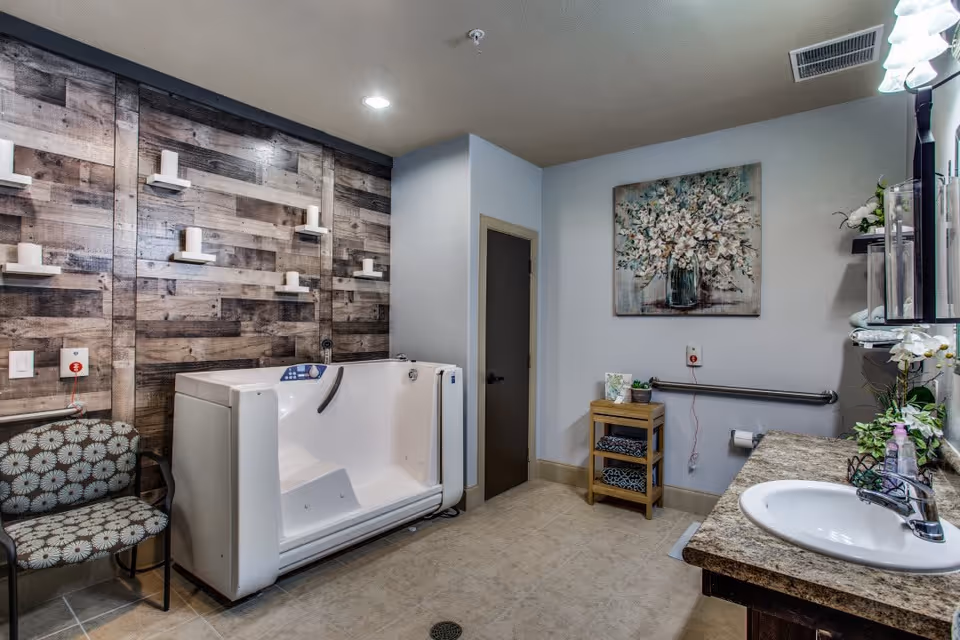 A spacious bathroom with a walk-in bathtub, a patterned chair, a wooden wall with small white shelves, a sink with a granite countertop, a small wooden shelf with towels, a floral painting on the wall, and various plants and toiletries.