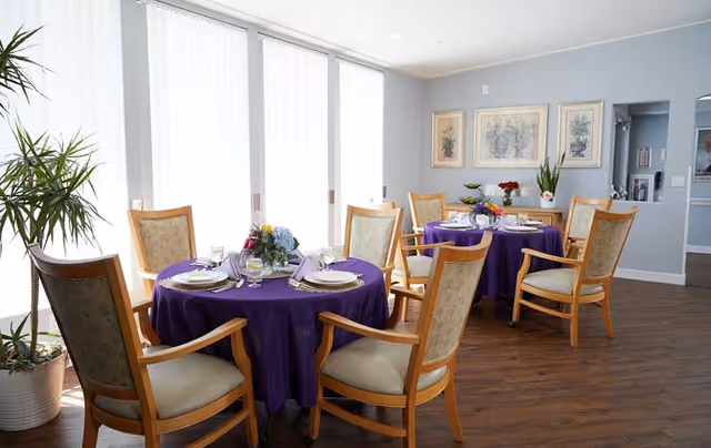 A bright dining room with two round tables covered in purple tablecloths, each set with plates, glasses, and floral centerpieces. The room has wooden chairs with cushioned seats and backs, large windows with white blinds letting in natural light, light blue walls with framed artwork, and a wooden floor.