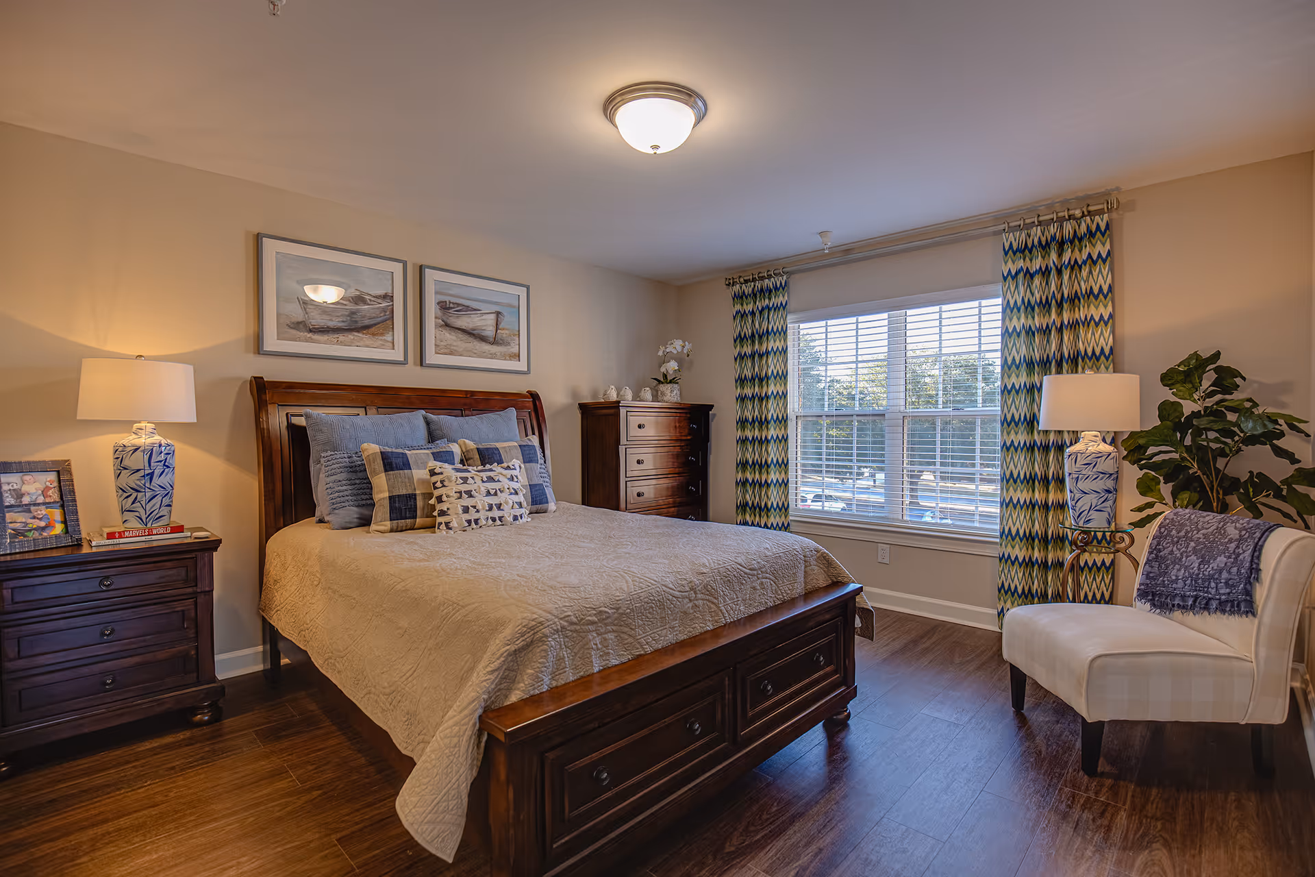 A cozy bedroom with a wooden bed frame and beige quilted bedspread, decorated with multiple pillows in blue and beige tones. Two framed pictures of boats hang above the bed. On the left side, there is a wooden nightstand with a blue and white lamp, a framed photo, and a book. On the right side, a white upholstered chair with a blue throw blanket sits next to a tall plant and another blue and white lamp. The room has wooden flooring, a large window with patterned blue and green curtains, and soft lighting from a ceiling fixture.