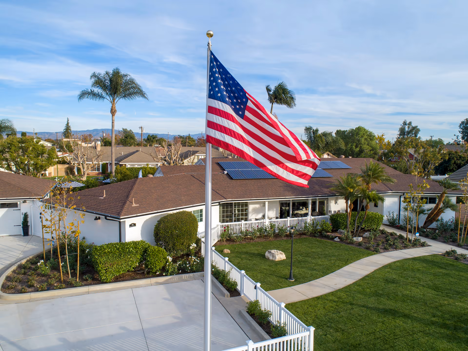 A single-story white building with a brown roof and solar panels, surrounded by a well-maintained garden with green grass, bushes, and palm trees. A large American flag on a flagpole is prominently displayed in the foreground. The sky is clear with some clouds, and there are mountains visible in the background.
