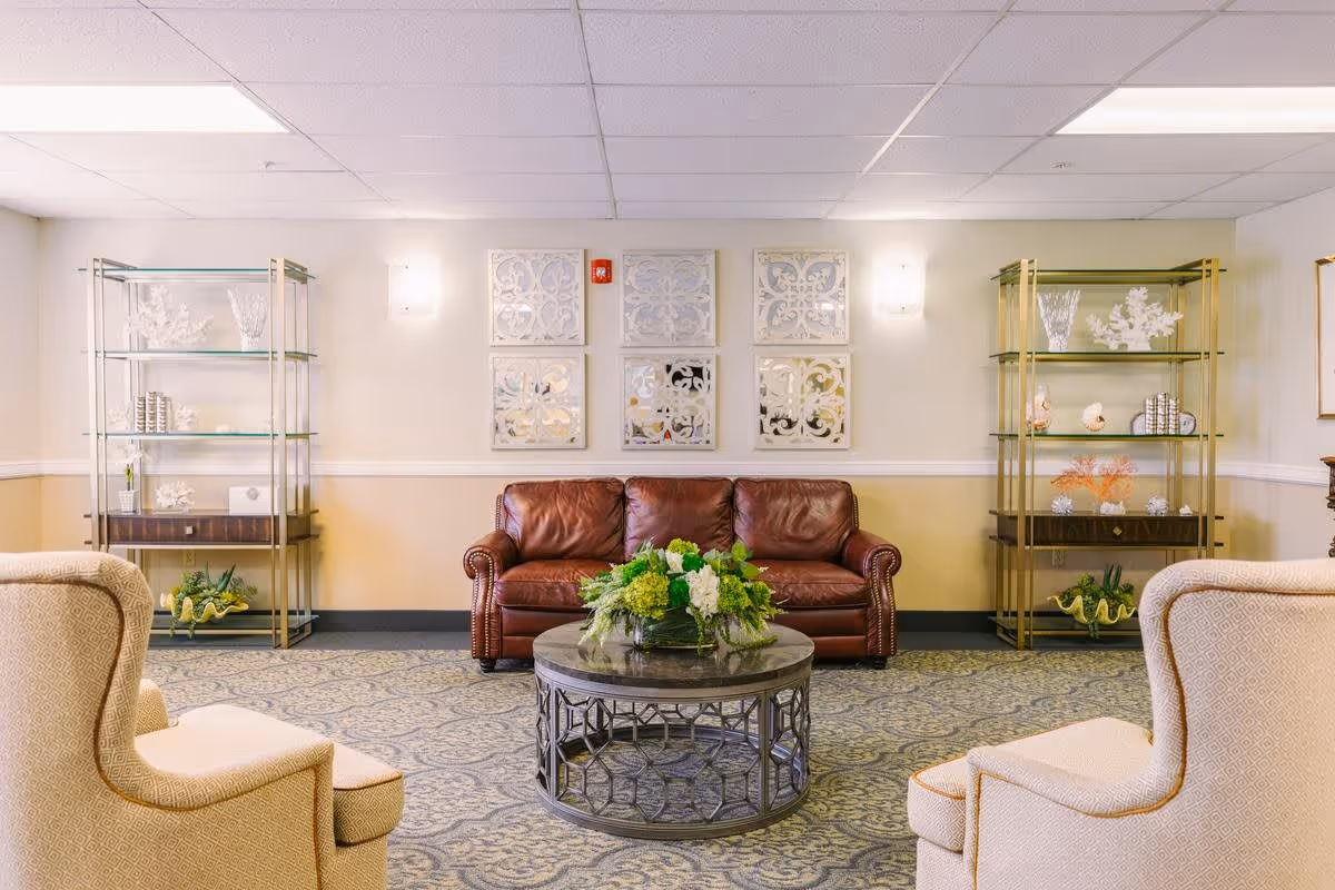A cozy senior living facility common area with a brown leather sofa centered against a wall adorned with six decorative square mirrors. Two beige upholstered armchairs face the sofa, with a round metal and glass coffee table holding a green floral arrangement in the middle. On either side of the sofa are tall glass and brass shelving units displaying decorative items including coral and books. The room has a patterned carpet and soft lighting from wall sconces.