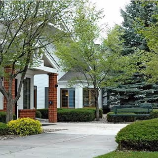 Landscaped front entrance of a senior living building with brick columns, white siding, trees, a bench, and a paved walkway.