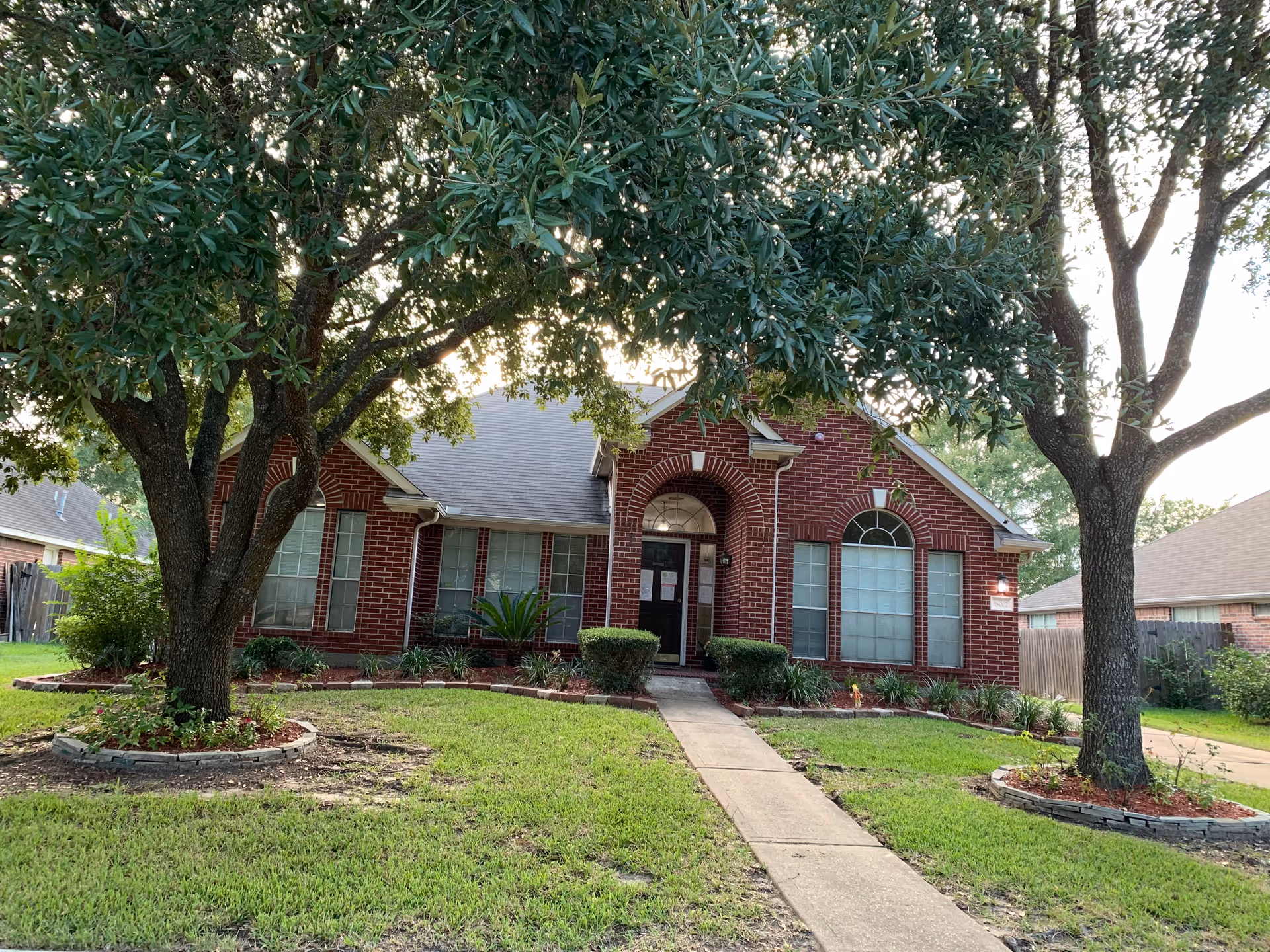 Brick single-story house with an arched front entry, large windows, a walkway, and two large trees in the front yard.