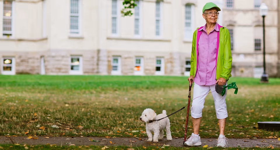 An elderly woman in bright clothing stands on a grassy lawn holding a leash attached to a small white dog in front of a large building.