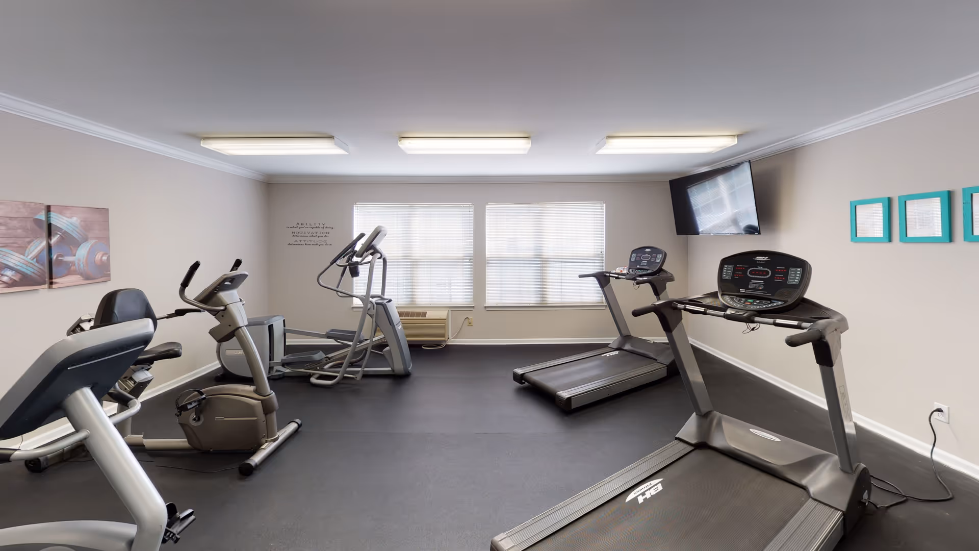 A small fitness room with exercise equipment including two treadmills, a stationary bike, and an elliptical machine. The room has beige walls, three fluorescent ceiling lights, a wall-mounted TV, and three windows with blinds. There are framed pictures on the walls and a motivational quote decal near the windows.