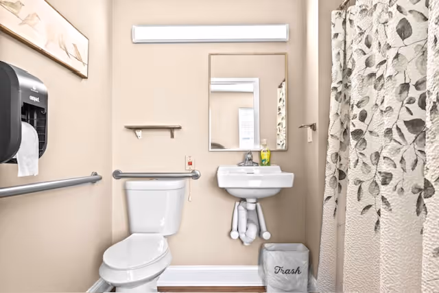 A small bathroom with beige walls featuring a white toilet, a white wall-mounted sink with a soap dispenser, a rectangular mirror above the sink, a trash bin labeled 'Trash', a shower with a curtain decorated with a leaf pattern, and a paper towel dispenser on the left wall.
