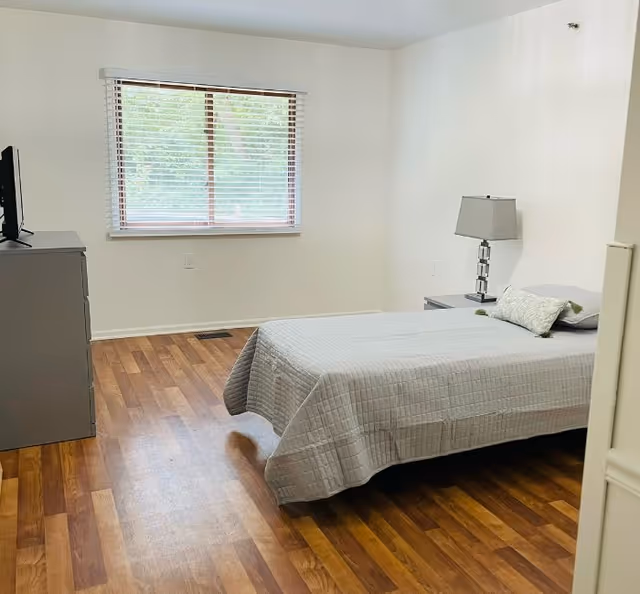 A simple bedroom with a single bed covered in a light gray quilt, a matching pillow, and a small decorative pillow. Next to the bed is a gray nightstand with a modern lamp. Across from the bed is a gray dresser with a TV on top. The room has wooden flooring and a window with white blinds letting in natural light.