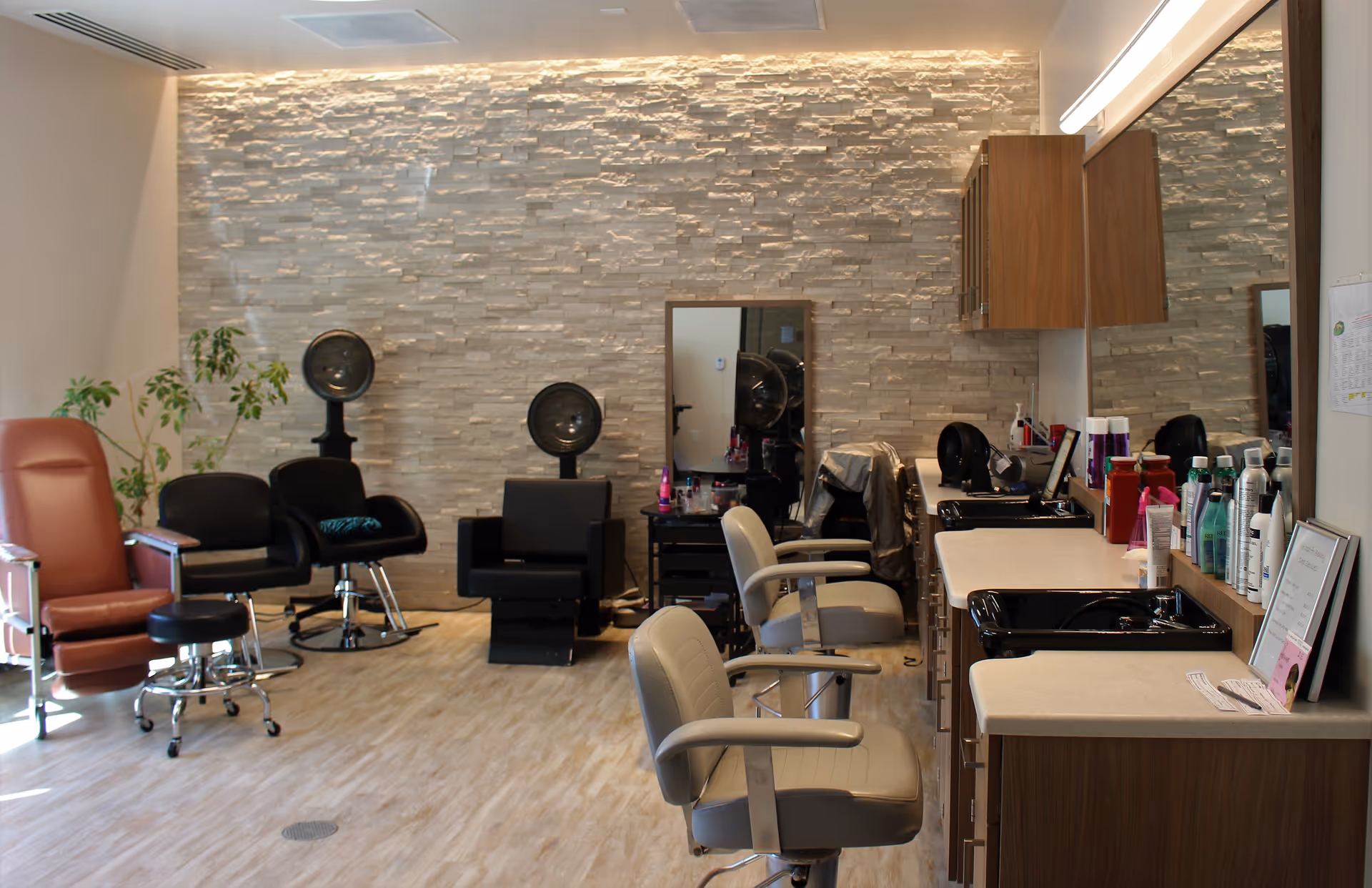Interior salon with styling chairs, hooded hair dryers, sinks, mirrors, and styling products against a stone accent wall.