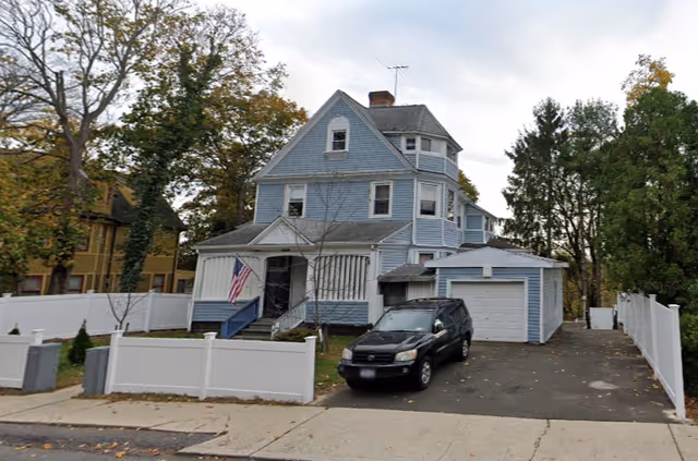 A three-story blue house with white trim and a small front porch displaying an American flag. There is a black SUV parked in the driveway next to a detached garage. The property is enclosed by a white fence and surrounded by trees with autumn foliage.