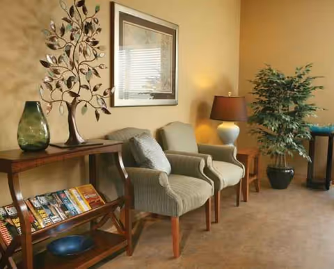 Cozy waiting lounge with three upholstered chairs, a plant, lamp, framed artwork, and magazines.