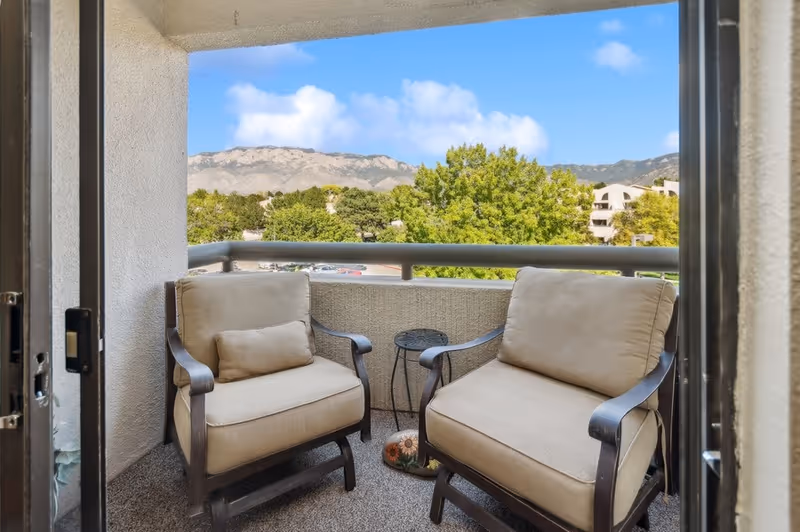 A small outdoor balcony with two cushioned armchairs and a small round metal table between them. The balcony overlooks a parking area, green trees, and distant mountains under a blue sky with some clouds.