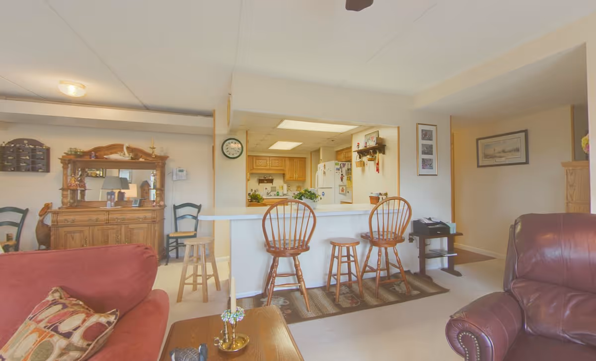 Interior view of a senior living facility showing a cozy living room with a red couch and a brown leather armchair. There is a wooden coffee table with decorative items in front of the couch. Behind the living room is a kitchen area with a white counter and four wooden stools. The kitchen has wooden cabinets and a white refrigerator. On the left side, there is a wooden cabinet with a mirror and decorative items on top. The walls are adorned with framed pictures and a clock.