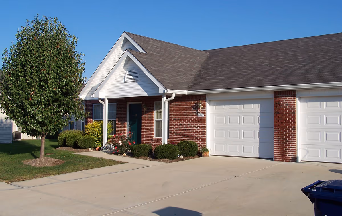 Front exterior of a single-story brick residence with attached garages, a small covered porch, and a tree by the driveway.