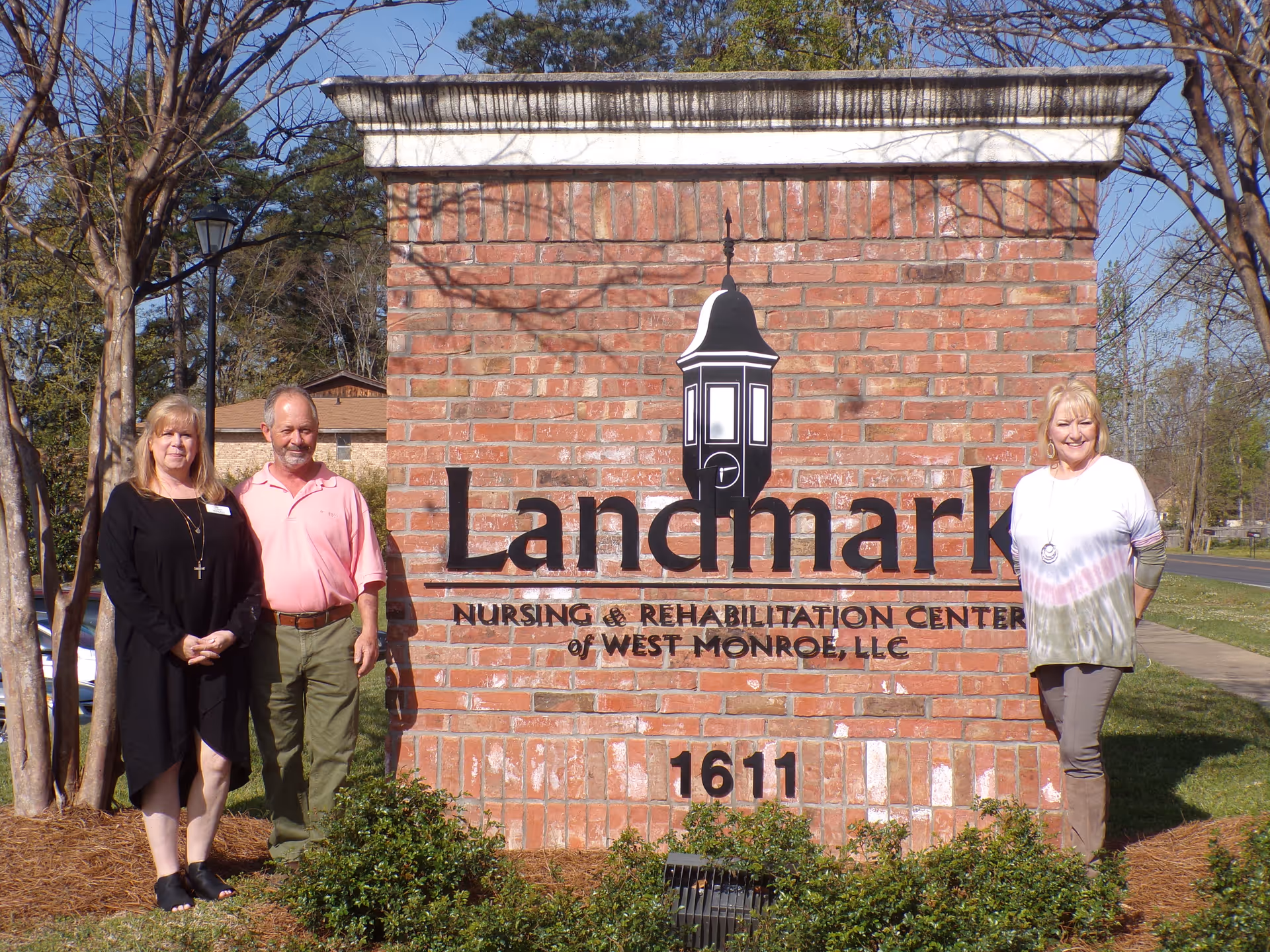Three people standing beside a large brick sign for Landmark Nursing & Rehabilitation Center of West Monroe, LLC.