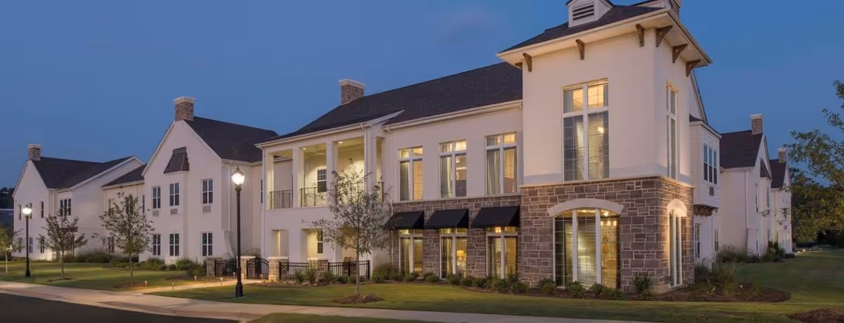 Exterior view of a large, multi-story senior living facility building at dusk with lights on inside. The building features a combination of stone and light-colored siding, multiple windows, balconies, and a well-maintained lawn with small trees and street lamps along the sidewalk.