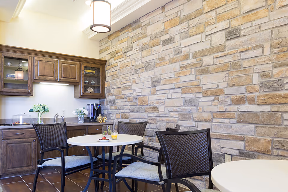 A cozy dining area with round white tables and black chairs with cushions. The background features a stone accent wall and wooden cabinets with glass doors. On the countertop, there are decorative flowers, a coffee maker, and a small plate with food and a glass of orange juice on one of the tables.