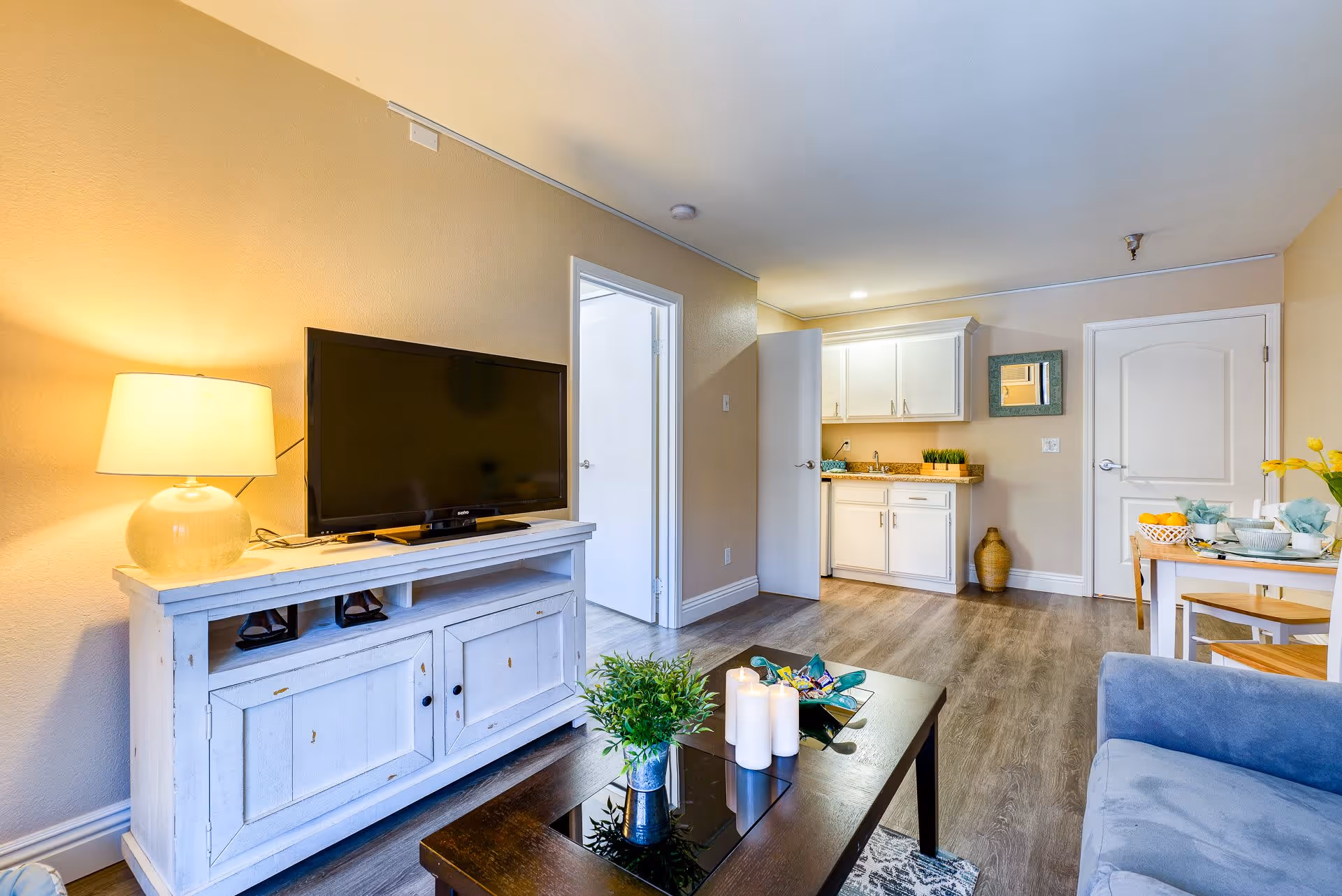 A bright and cozy senior living interior featuring a white TV stand with a flat-screen TV and a lamp on the left. In the foreground, there is a dark wooden coffee table with candles and a small plant. To the right, a blue sofa is partially visible. The background shows a small kitchenette with white cabinets, a countertop, and a decorative vase on the floor. A dining table set with plates, bowls, and yellow flowers is also visible near a closed white door.