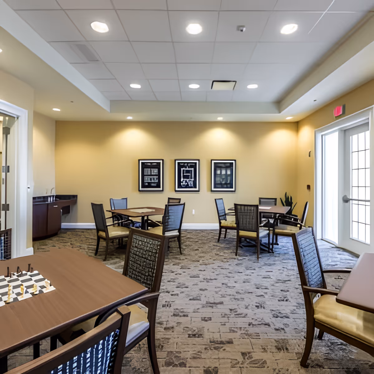 Well-lit communal dining/activity room with multiple tables and chairs, framed artwork on a yellow wall, and glass doors to the outside.