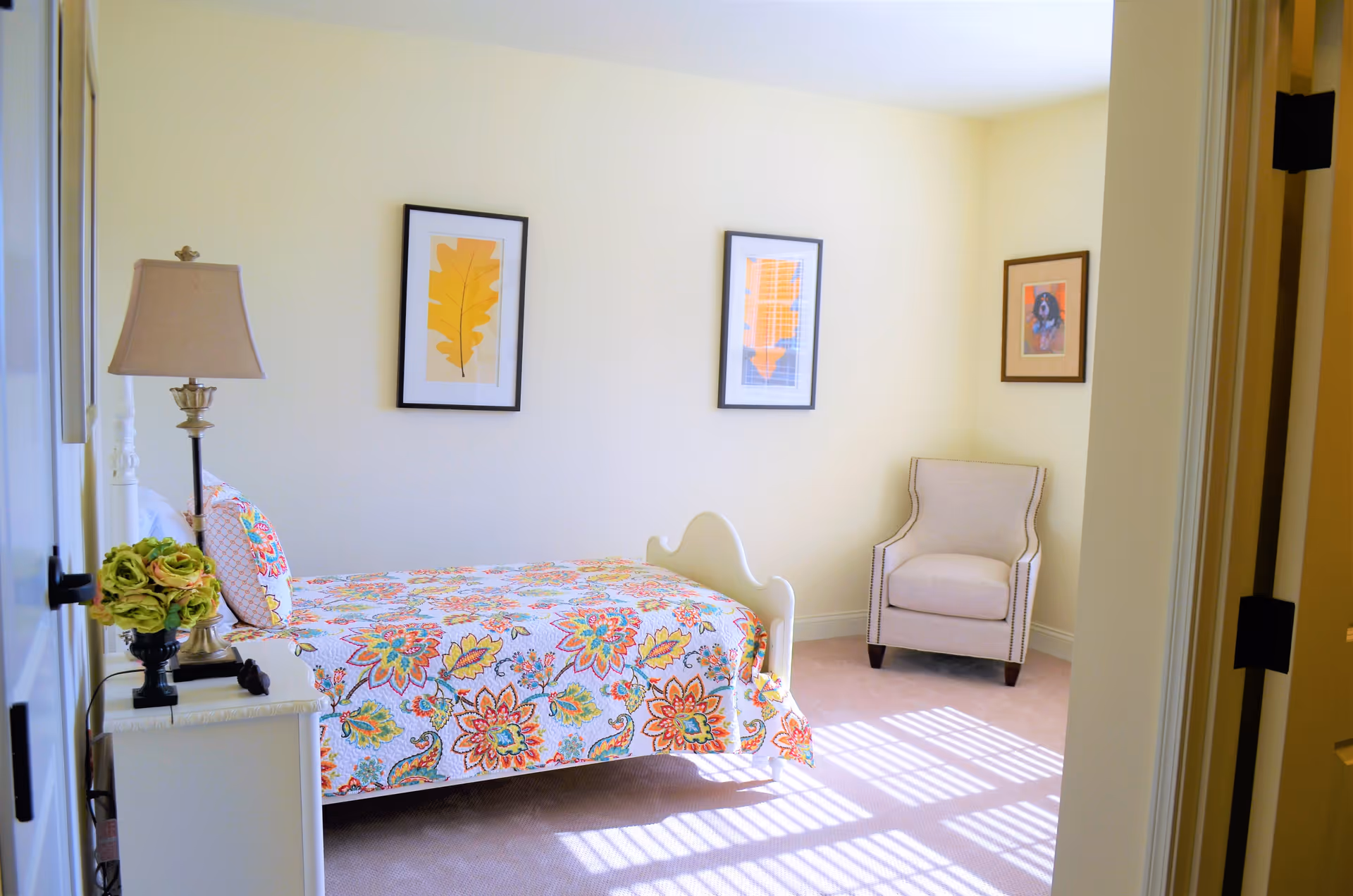A bright bedroom with a single bed covered in a colorful floral quilt. There is a white nightstand with a lamp and a small plant next to the bed. Three framed pictures hang on the light yellow walls, and a beige armchair is positioned in the corner near the window, which casts sunlight patterns on the carpeted floor.