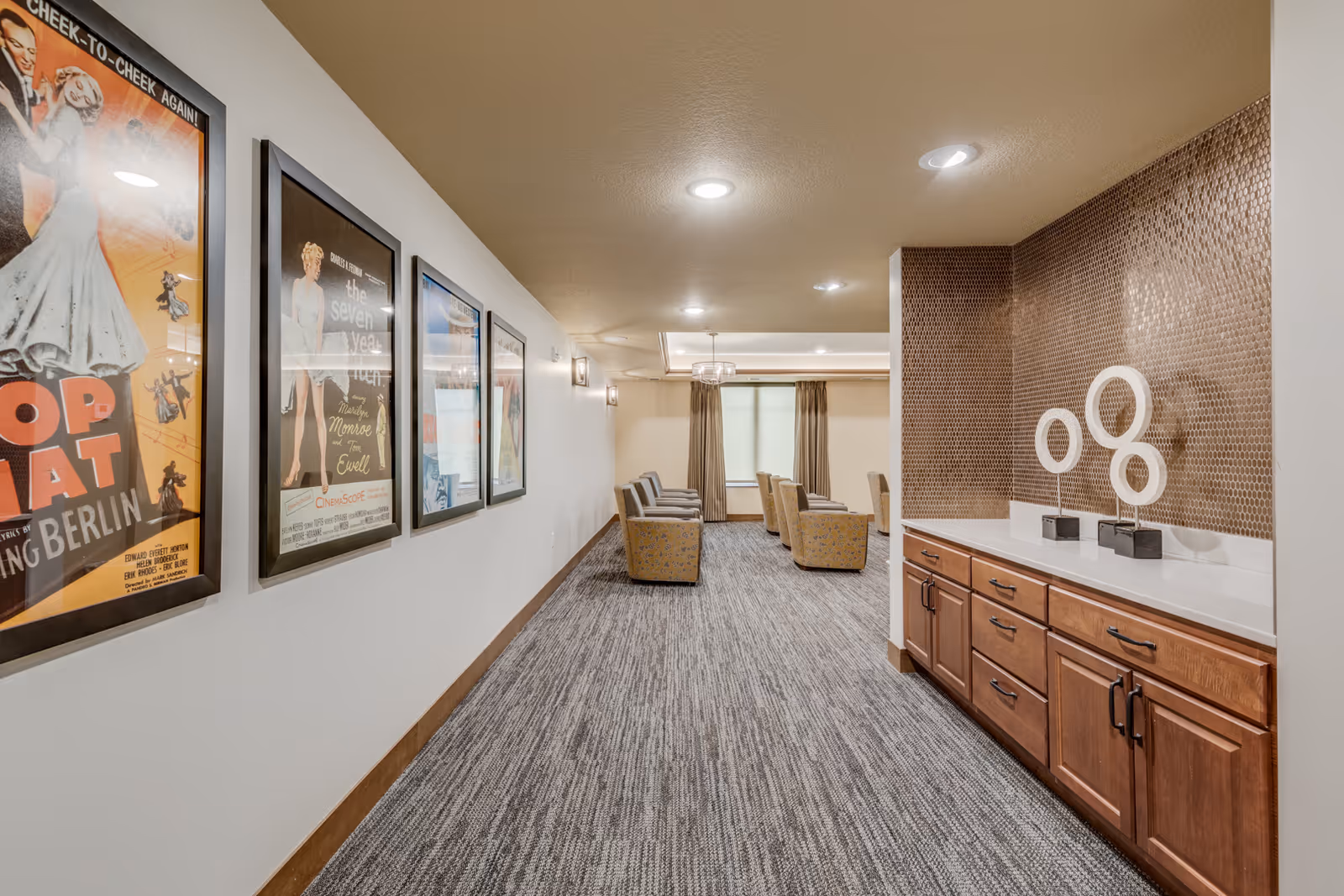 Carpeted hallway leading to a seating lounge with framed movie posters on the left and a countertop with wooden cabinets and decorative sculptures on the right.
