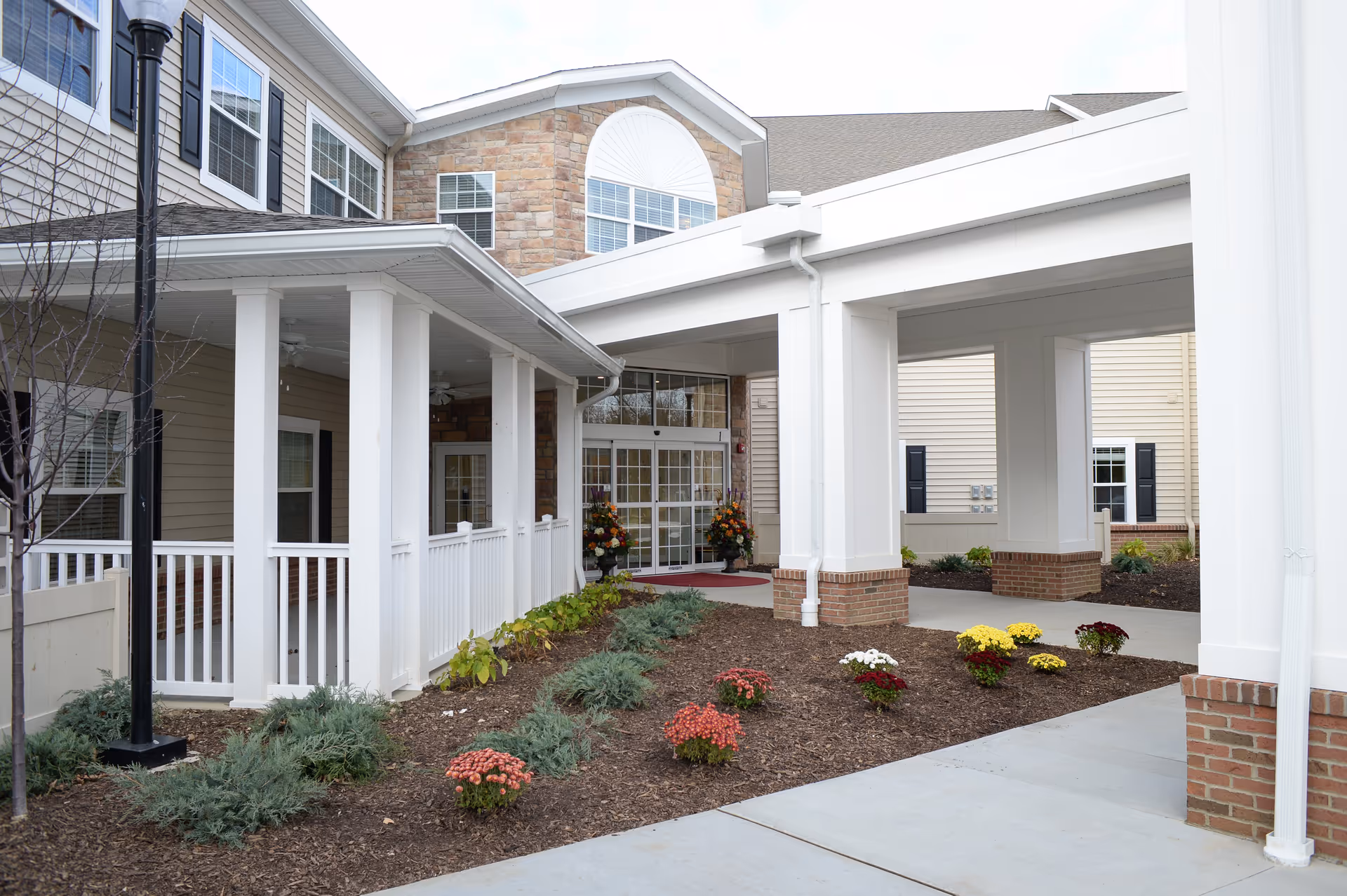 Covered entrance and porte-cochère of a senior living building with white columns, landscaping, and glass double doors.