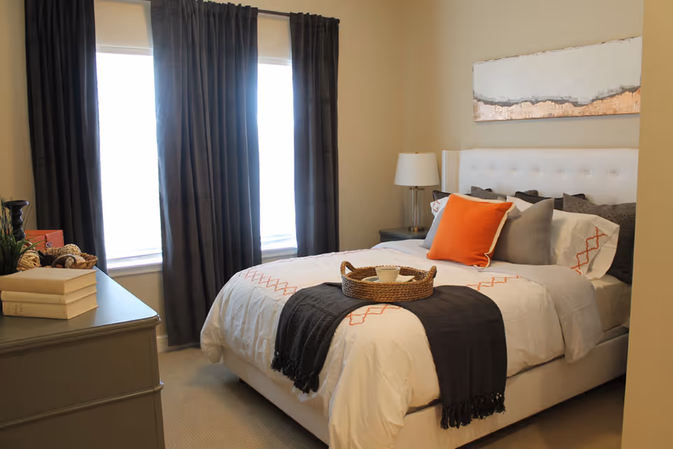 A cozy bedroom with a white upholstered bed dressed in white and orange linens, dark curtains over two windows, a bedside table with a lamp, and a dresser with books.