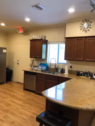 Interior view of a kitchen area with wooden cabinets, a granite countertop with a bar stool, a stainless steel dishwasher, a sink under a window, and a refrigerator. The word HOPE is displayed on top of one cabinet, and a modern clock is mounted on the wall. The floor is wooden, and there is an exit door in the background.
