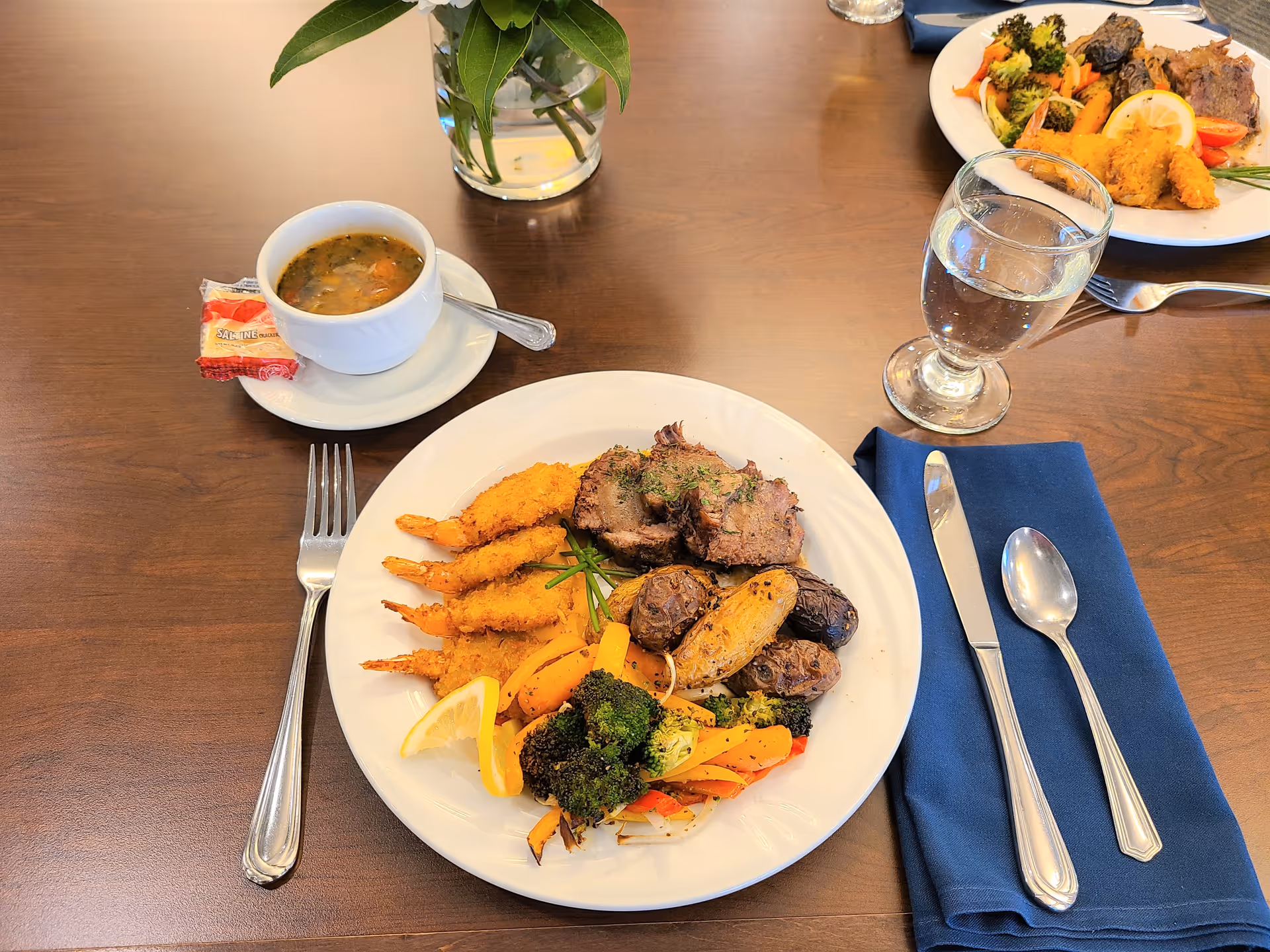 A dining table set with a plate of food including breaded shrimp, sliced meat, roasted potatoes, and mixed vegetables with a lemon wedge. There is a bowl of soup with a spoon on a saucer and a packet of crackers next to it. A glass of water and silverware on a blue napkin are also on the table. Another similar plate is partially visible in the background.
